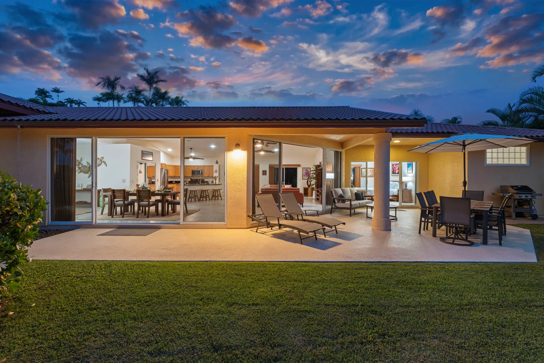 A covered patio with outdoor seating faces a brightly lit house at dusk.