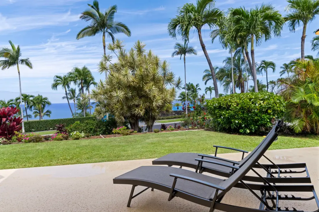 Outdoor lounge chairs sit on a paved patio beside a tropical garden.