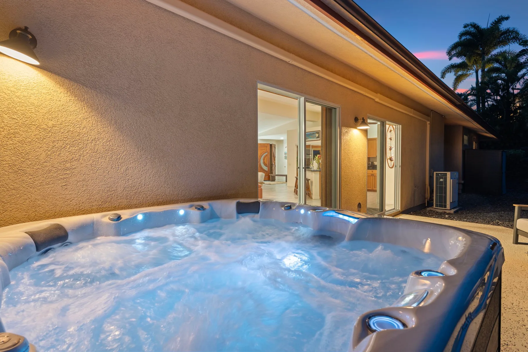 An outdoor spa sits beside a house, with illuminated windows and bubbling water.