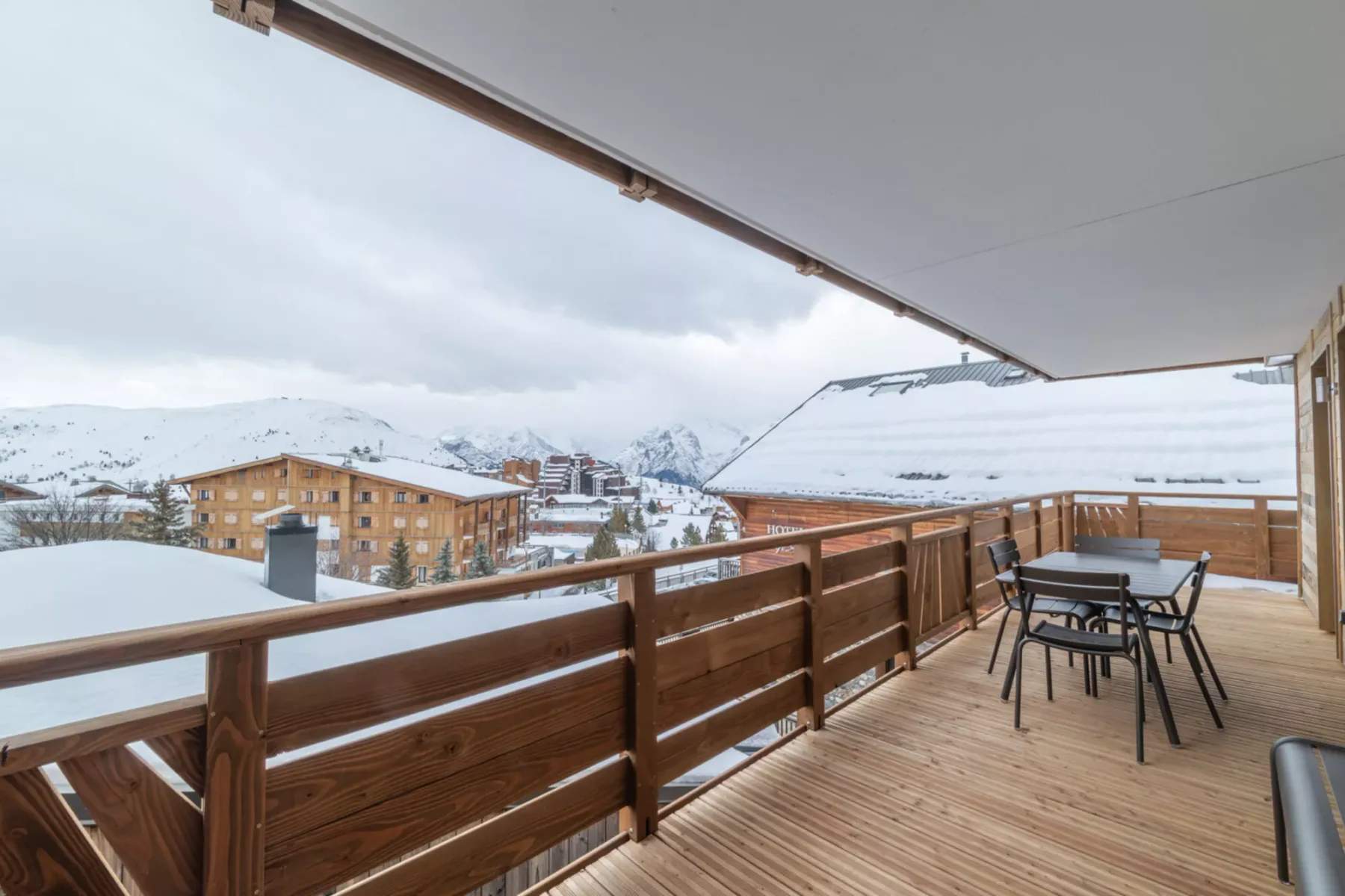 A covered wooden balcony with outdoor seating overlooks snow-covered buildings and mountains.