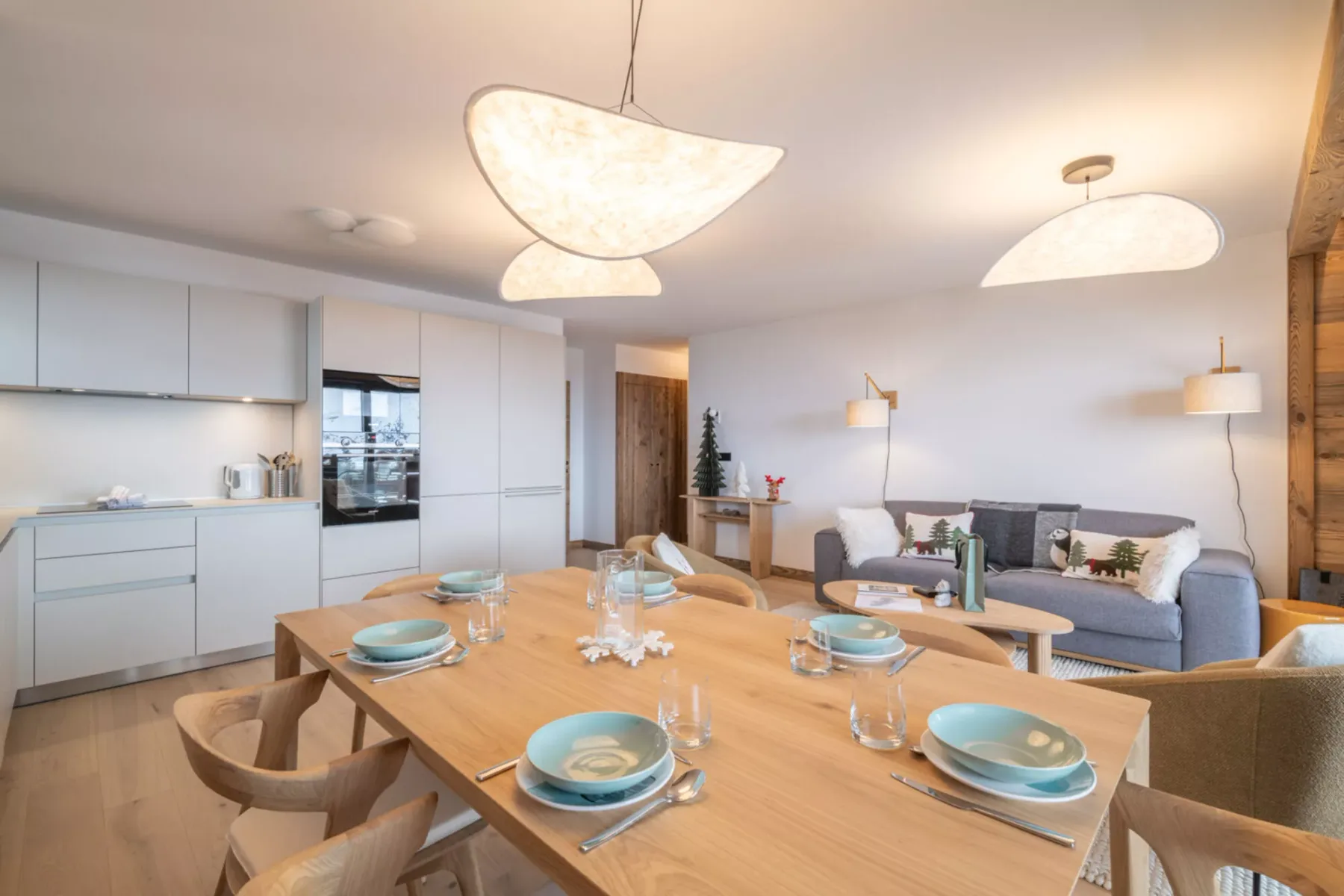 A bright open-plan kitchen and living area has a wooden dining table in the foreground.