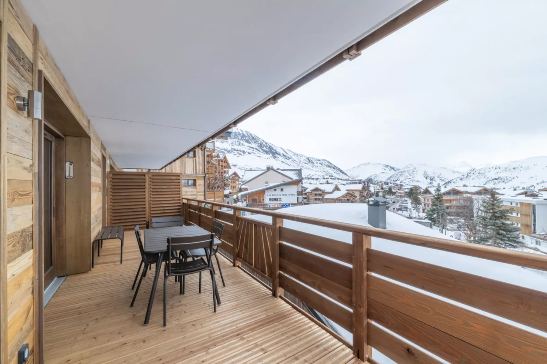 A covered wooden balcony has outdoor seating and overlooks snow-covered mountains.
