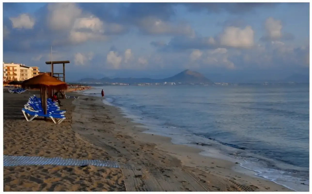 Una playa de arena junto a aguas tranquilas, con asientos y montañas a lo lejos.