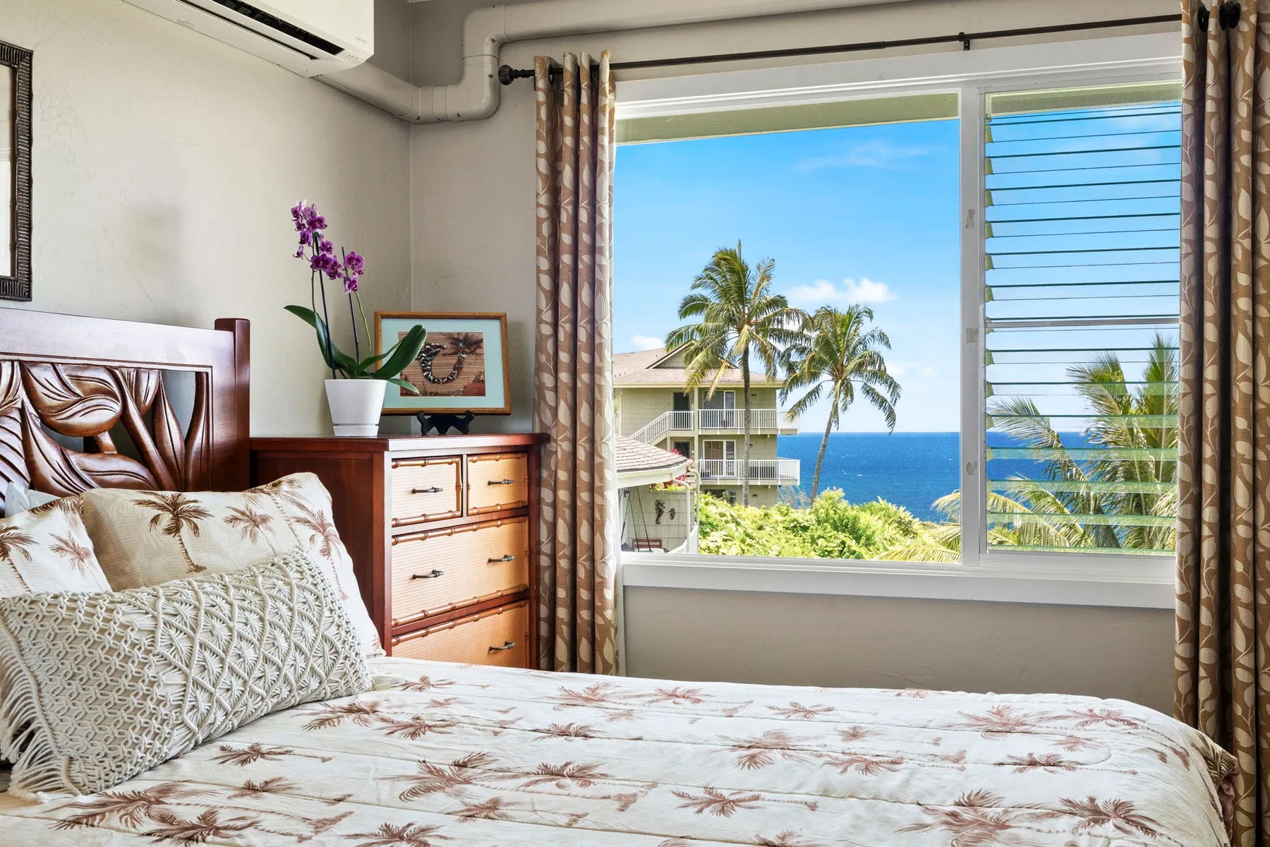 Un dormitorio con una colcha estampada y una ventana amplia con vistas a palmeras y al océano.