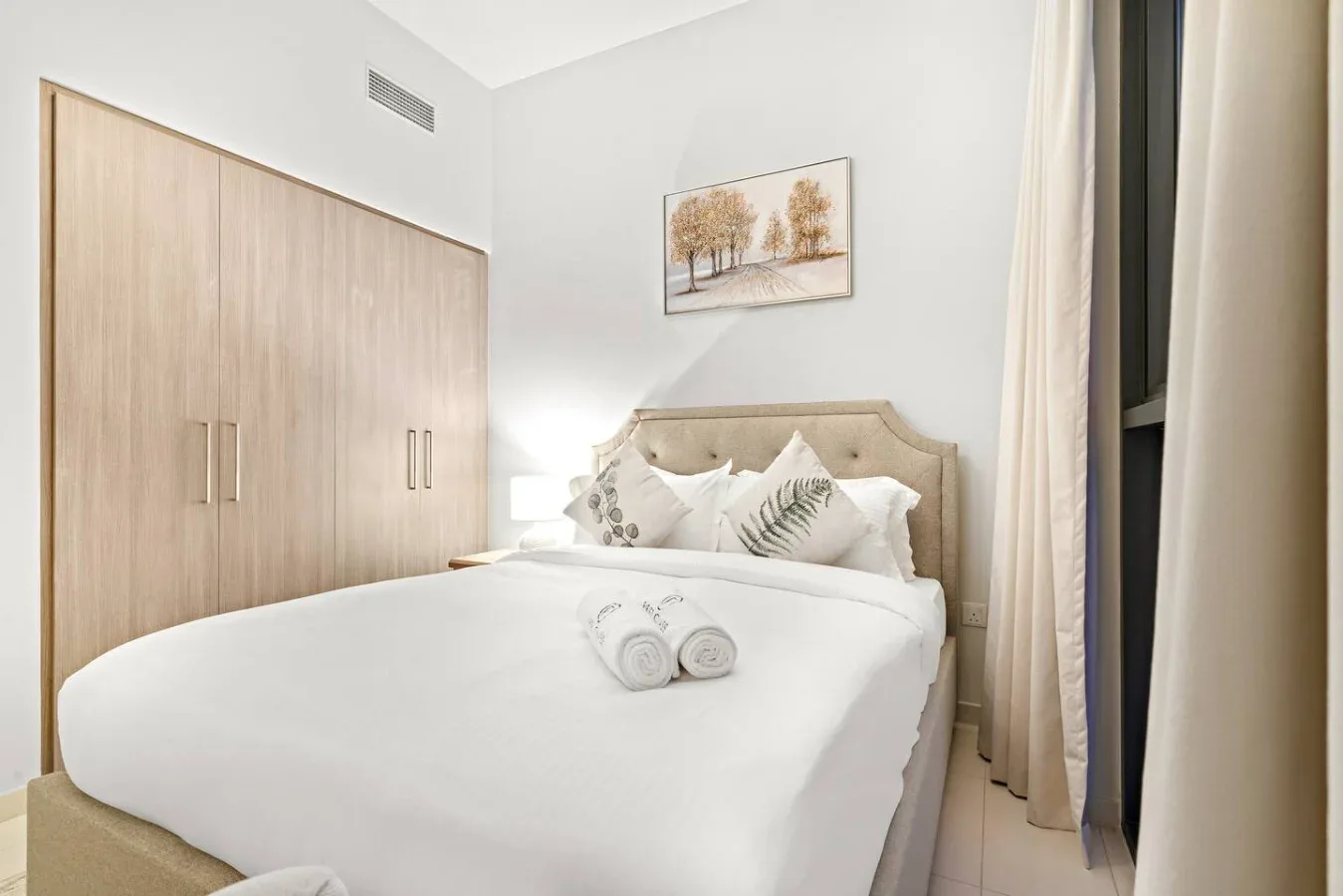 A bright bedroom with a neatly made bed and a framed landscape above the headboard.