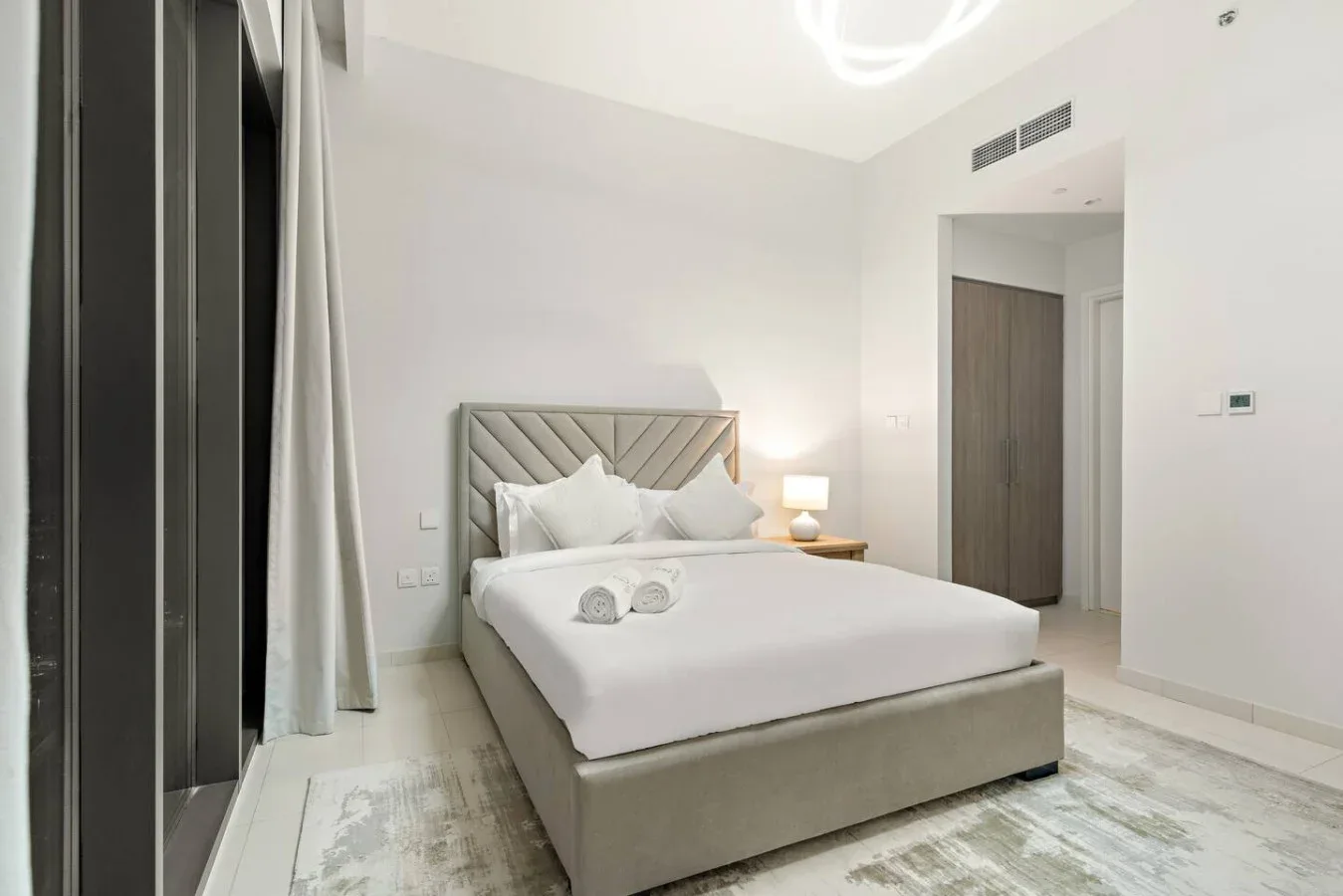 A bright bedroom with a neatly made bed and pale flooring.