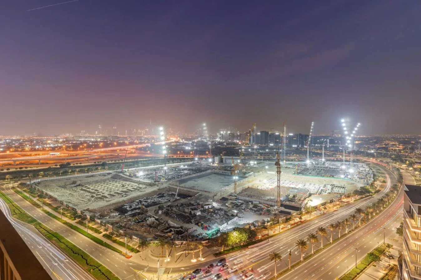 A large construction site at night is surrounded by illuminated roads and city lights.