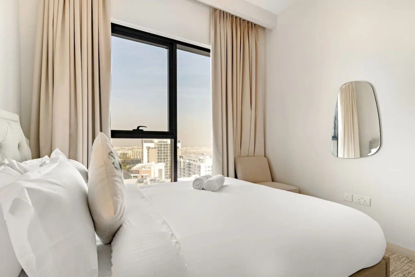 A bright bedroom with white bedding, beige curtains, and a large window.