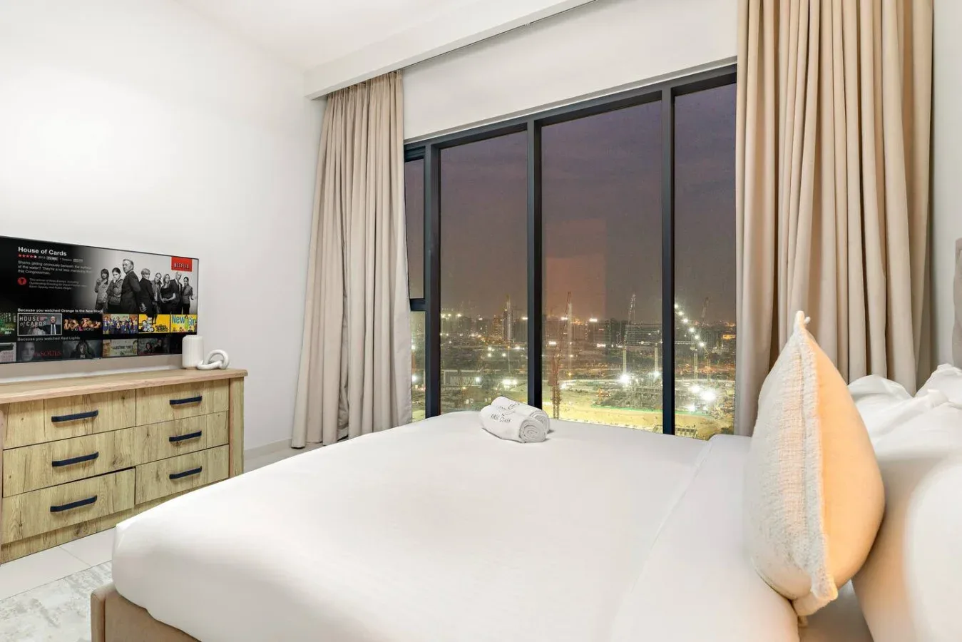 A bedroom with a bed beside large windows overlooking city lights.