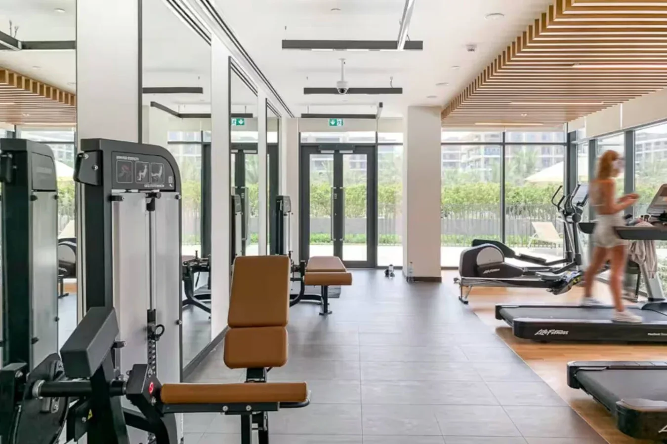A bright gym with exercise machines and treadmills beside large windows.