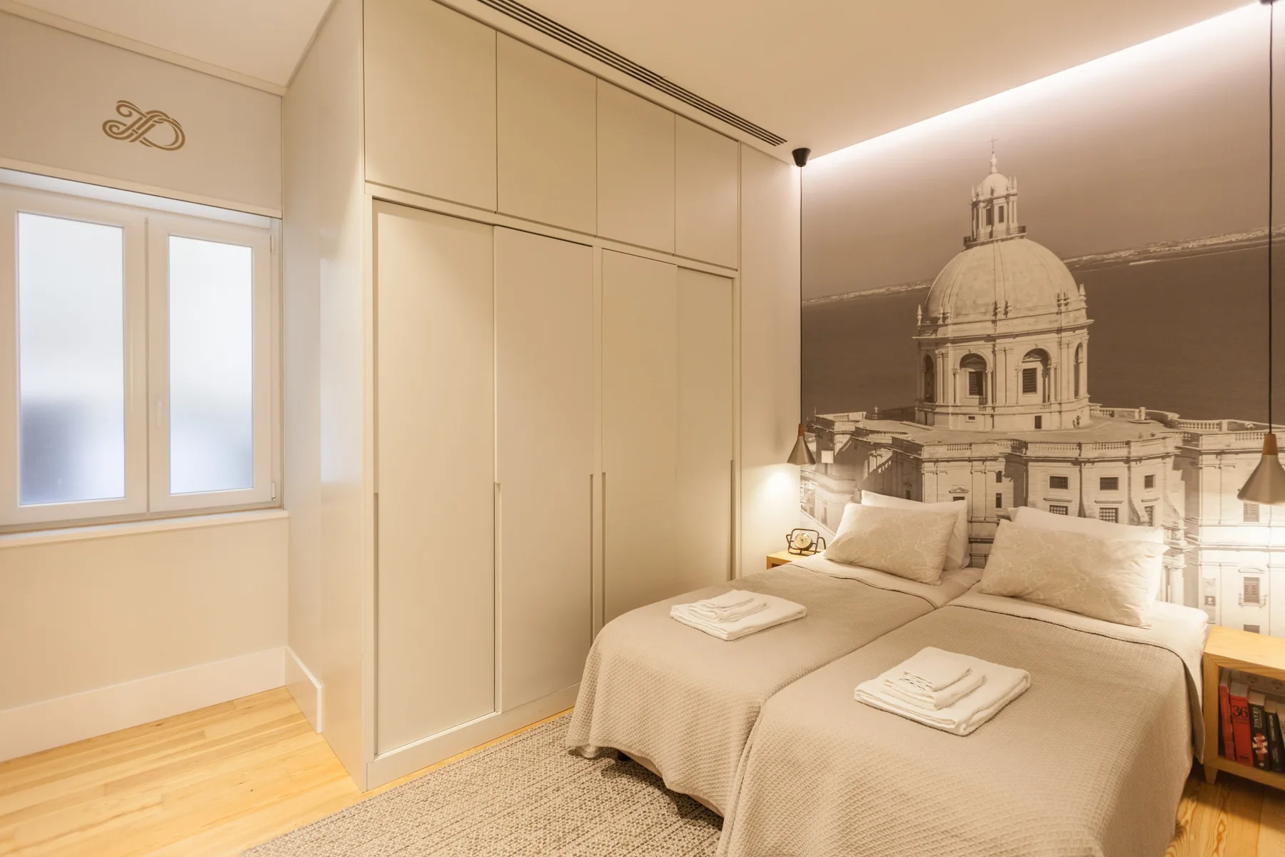 A bedroom with two beds, a built-in wardrobe, and a large architectural wall mural.
