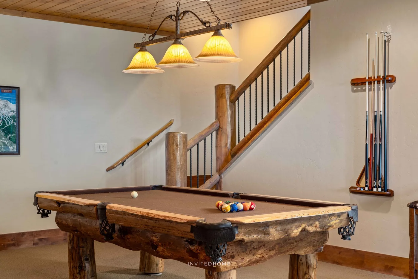 Una mesa de billar de madera está bajo una escalera en una sala de juegos bien iluminada.