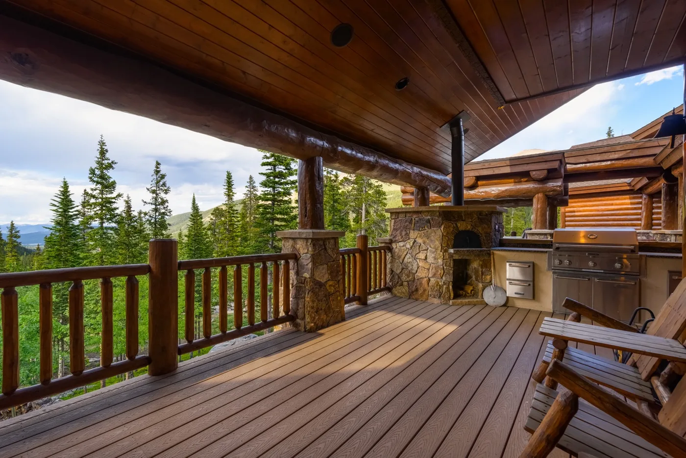 Una terraza de madera cubierta da a árboles de hoja perenne, con una chimenea de piedra y cocina exterior.