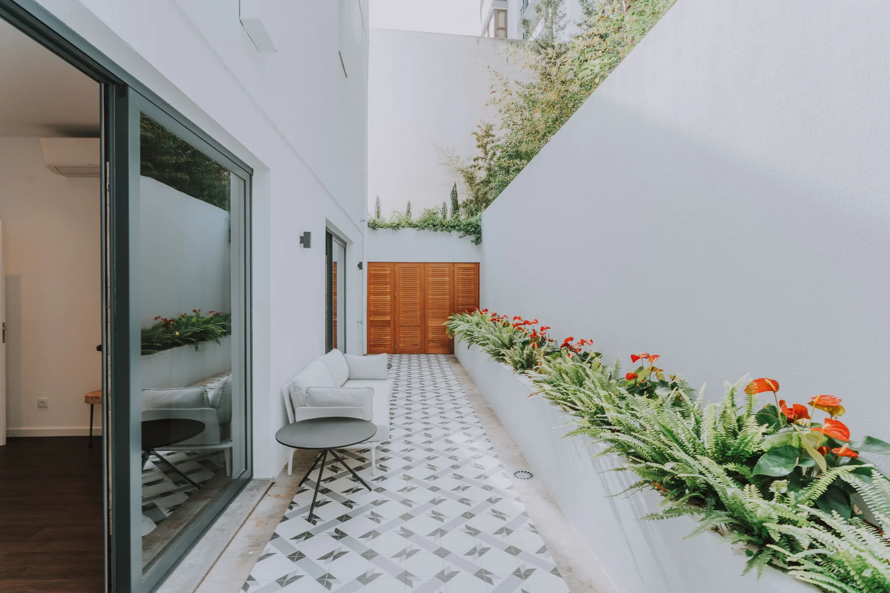 Un patio exterior con suelo de baldosas, asientos y un largo parterre junto al edificio.