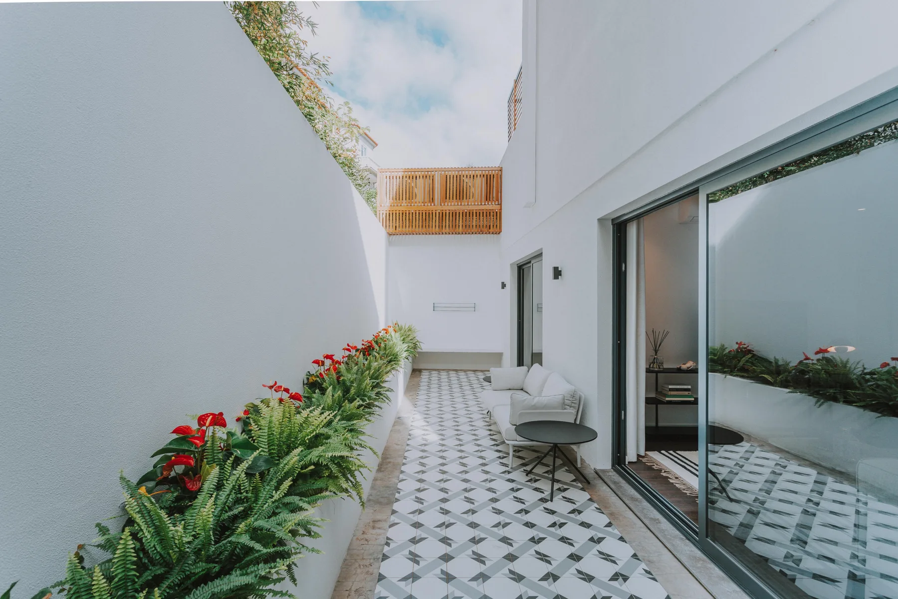 Un patio exterior con suelo de baldosas, plantas en macetas y asientos junto a una pared de cristal.
