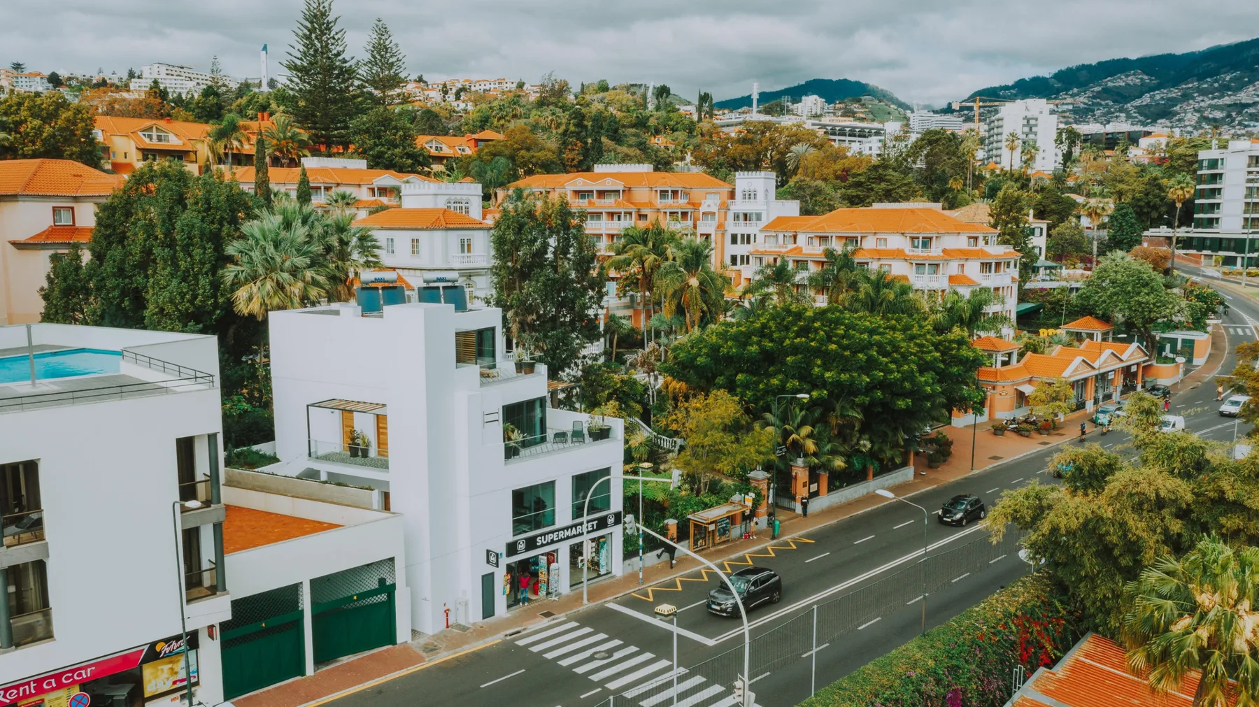 Una calle urbana bordeada de edificios y árboles, con colinas visibles al fondo.