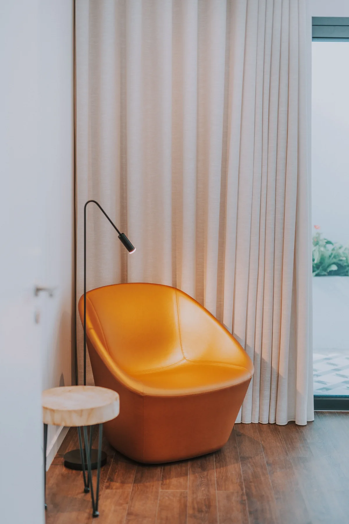Un asiento naranja brillante está junto a una lámpara de pie y una mesita, frente a cortinas altas.