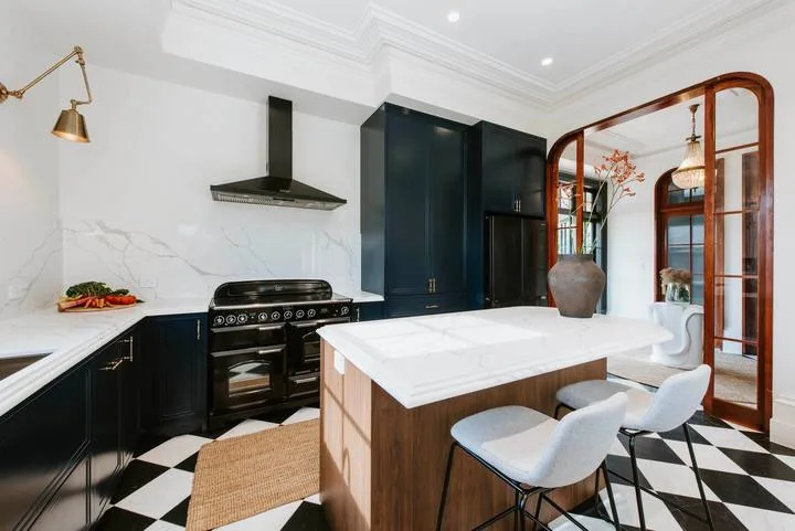 Una cocina luminosa con isla central, gabinetes oscuros y suelo de baldosas blancas y negras.