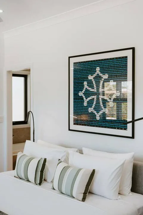 Un dormitorio con una cama blanca y cojines a rayas bajo un cuadro enmarcado.
