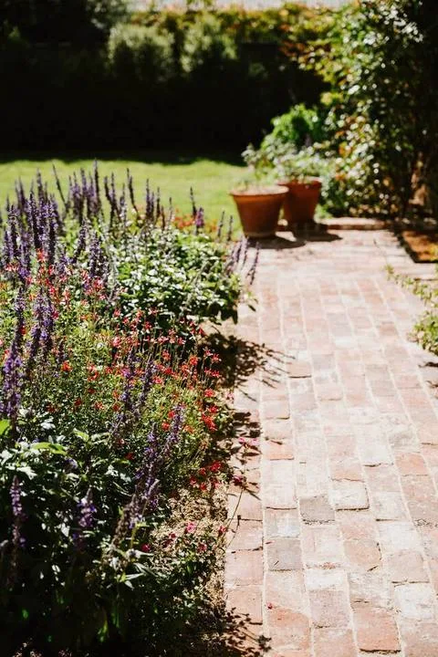 Un sendero de ladrillo bordea parterres de flores coloridas y plantas en macetas.