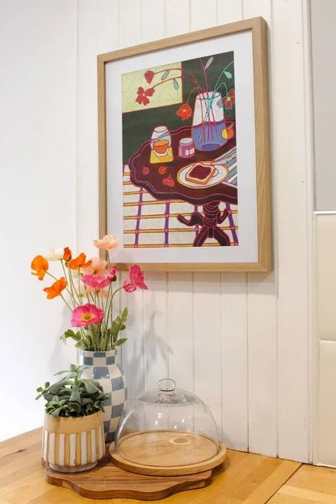 Una pintura colorida enmarcada cuelga sobre plantas en macetas y una vitrina de vidrio para pasteles.