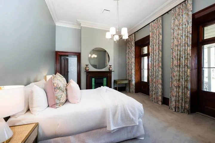 Un dormitorio con una cama blanca, cortinas estampadas y ventanas altas.