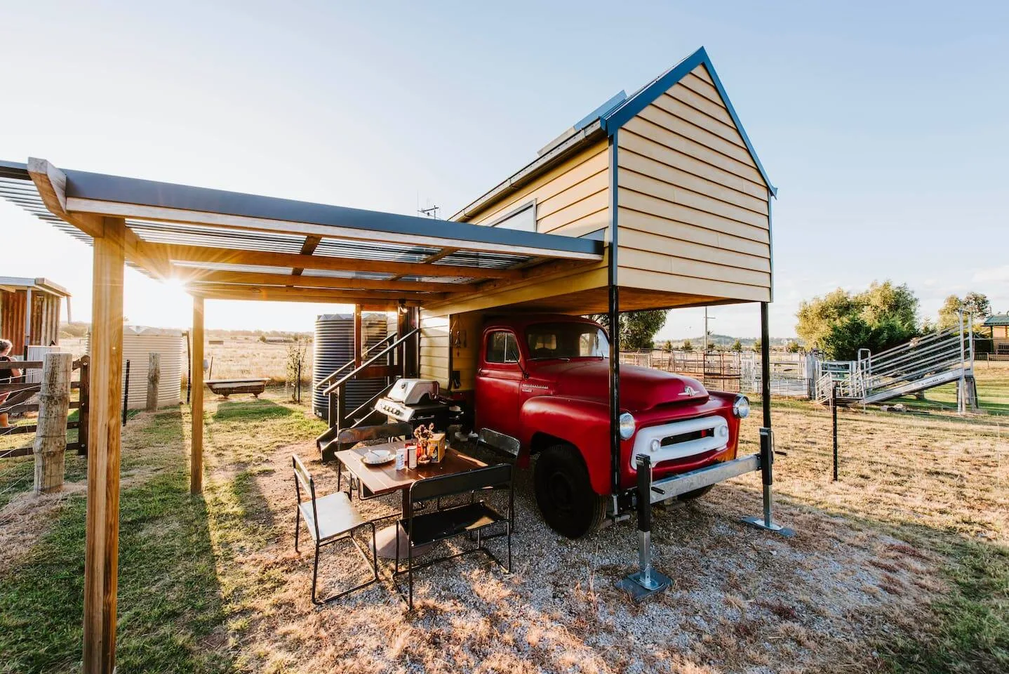 Una zona de comedor exterior cubierta está junto a una camioneta roja antigua en un entorno rural.