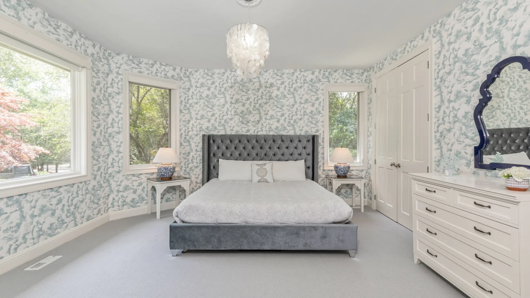 Un dormitorio con una cama, papel tapiz estampado y ventanas con vista a los árboles.