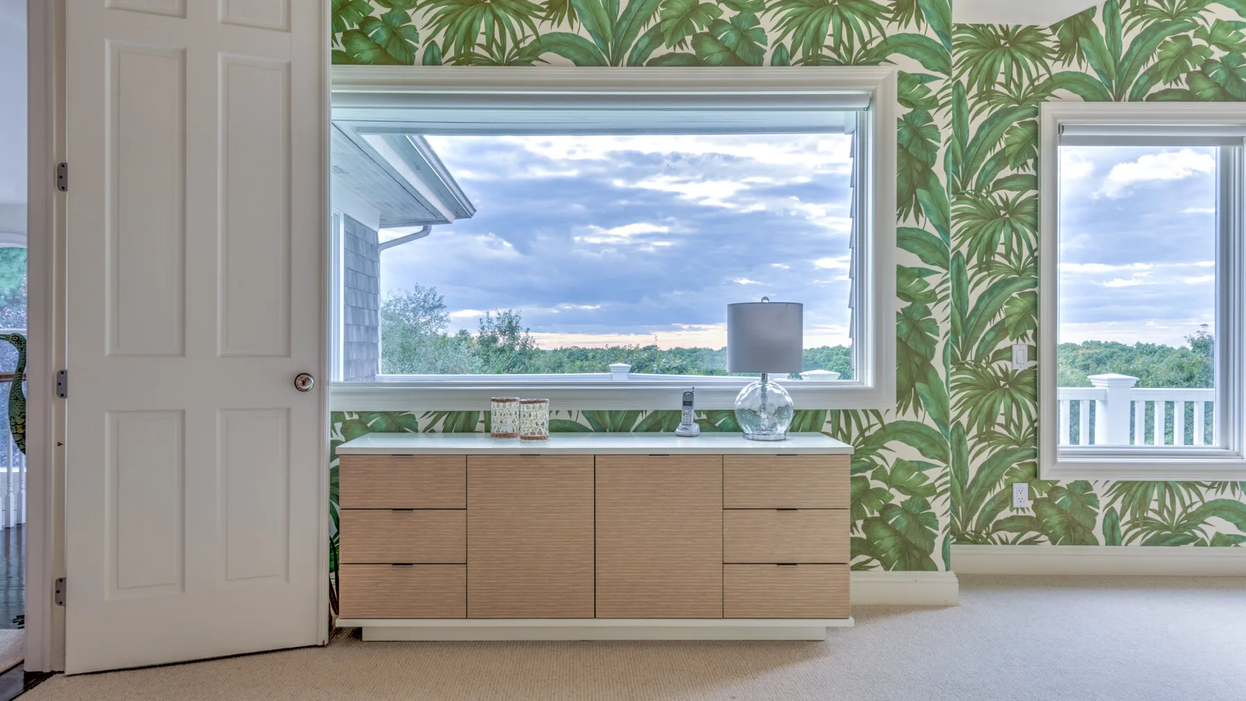 Una habitación con papel tapiz de hojas, un mueble bajo y ventanas con vistas a la vegetación.