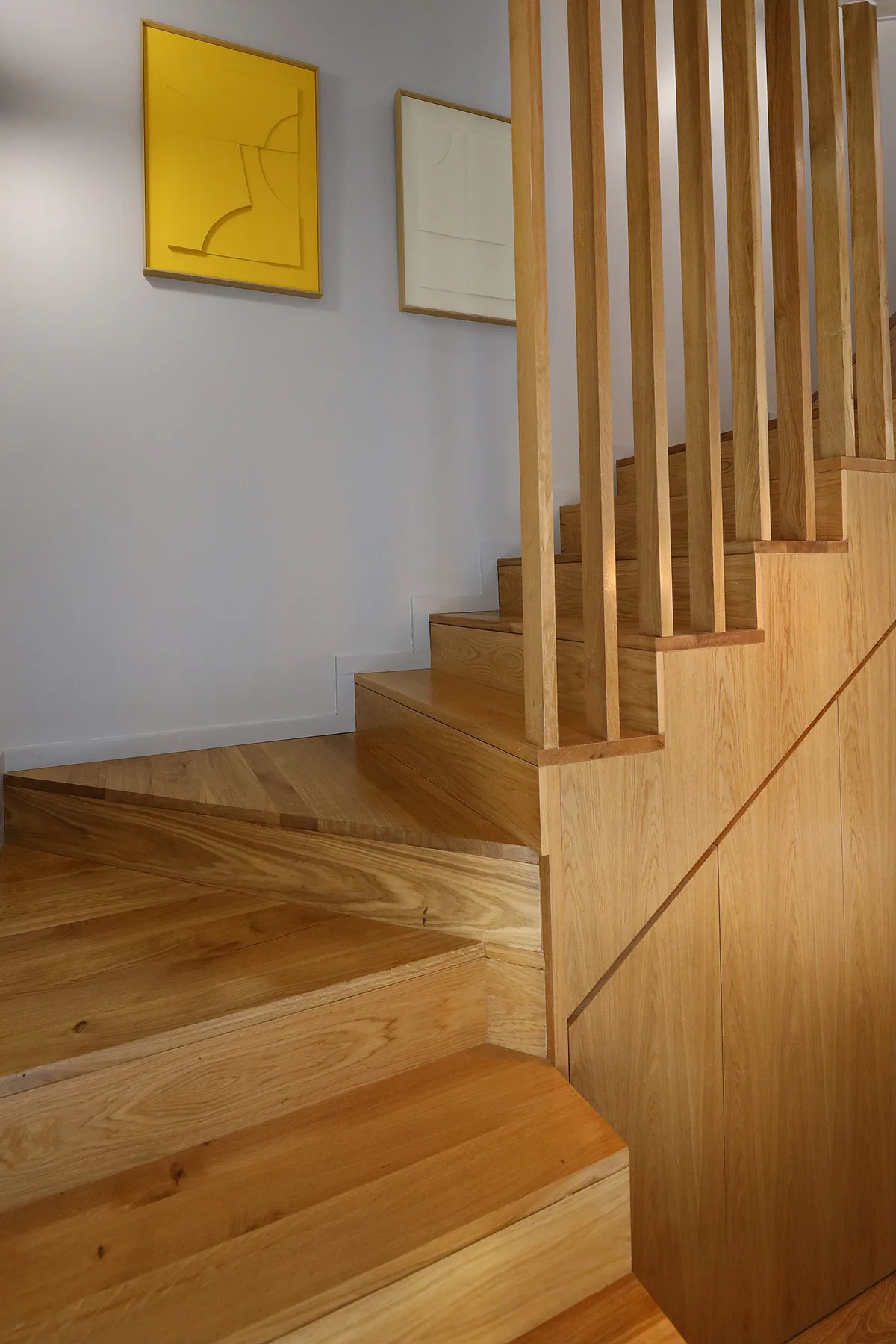A wooden staircase with vertical slats rises beside framed artwork on a white wall.