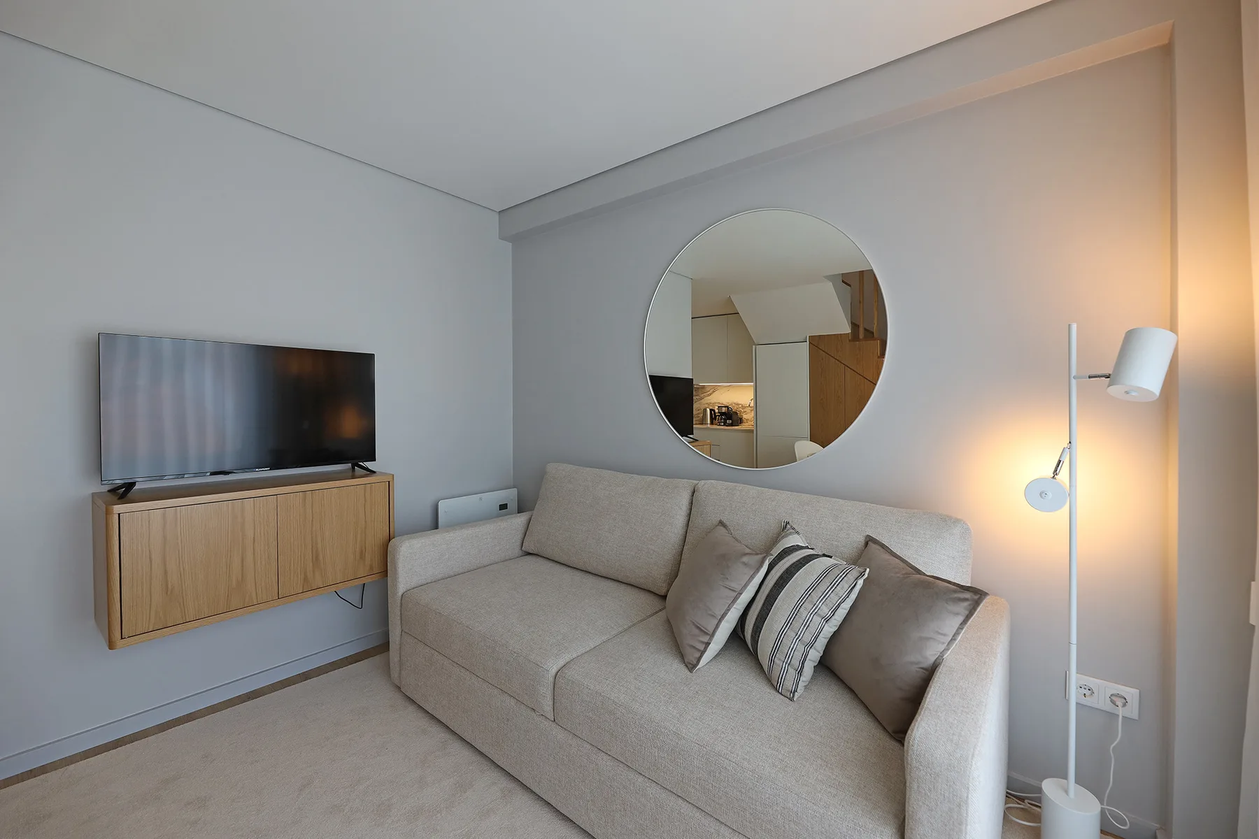 A living room with a beige sofa, wall-mounted television, and round mirror.