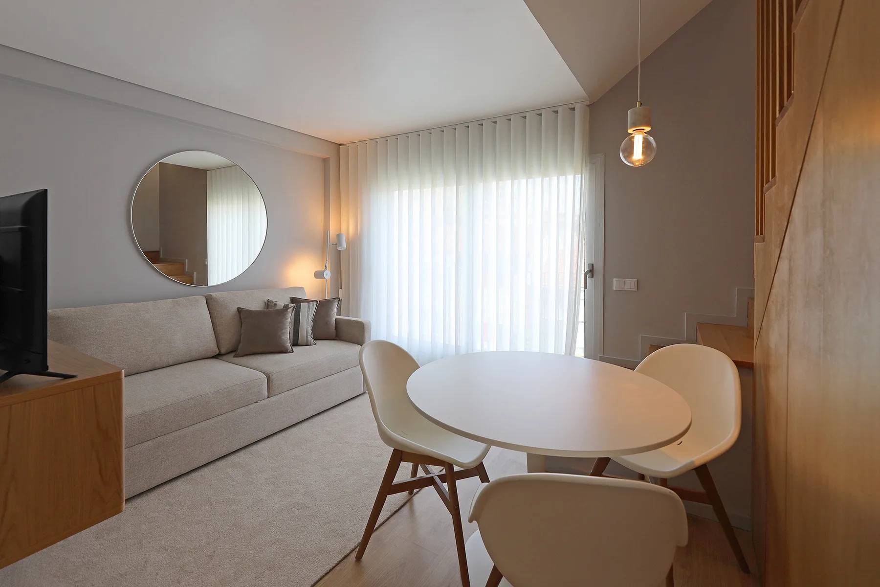 A bright living and dining area with a sofa, round table, and large curtained window.