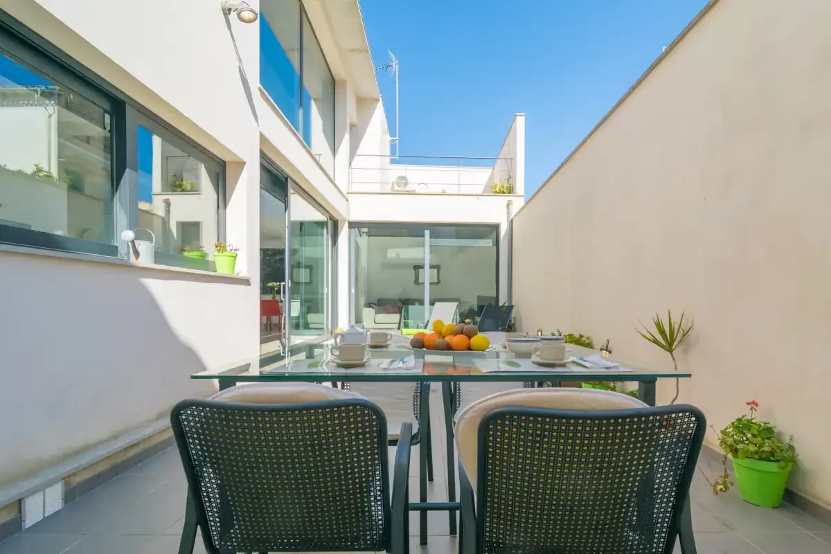 An outdoor dining area sits between a modern house and a tall wall.
