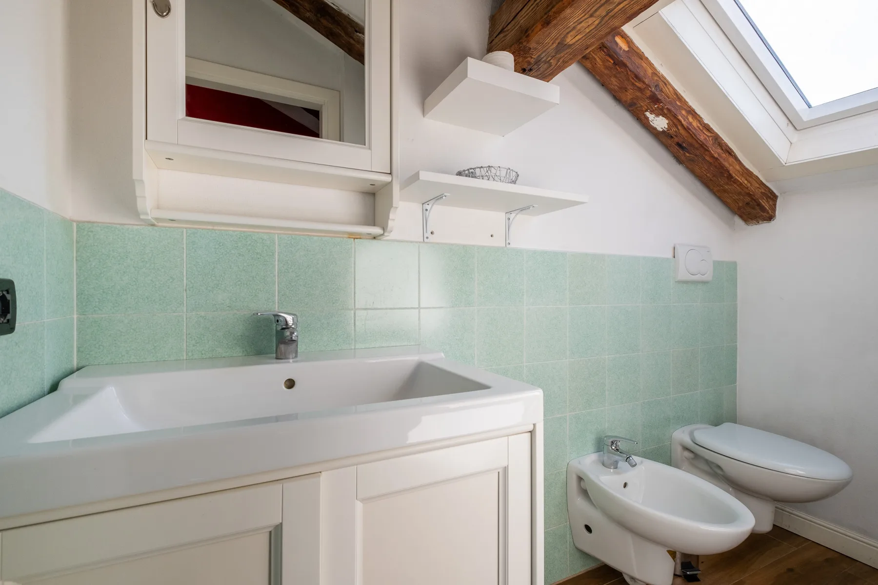 Un baño alicatado con lavabo, bidé e inodoro bajo un techo inclinado.