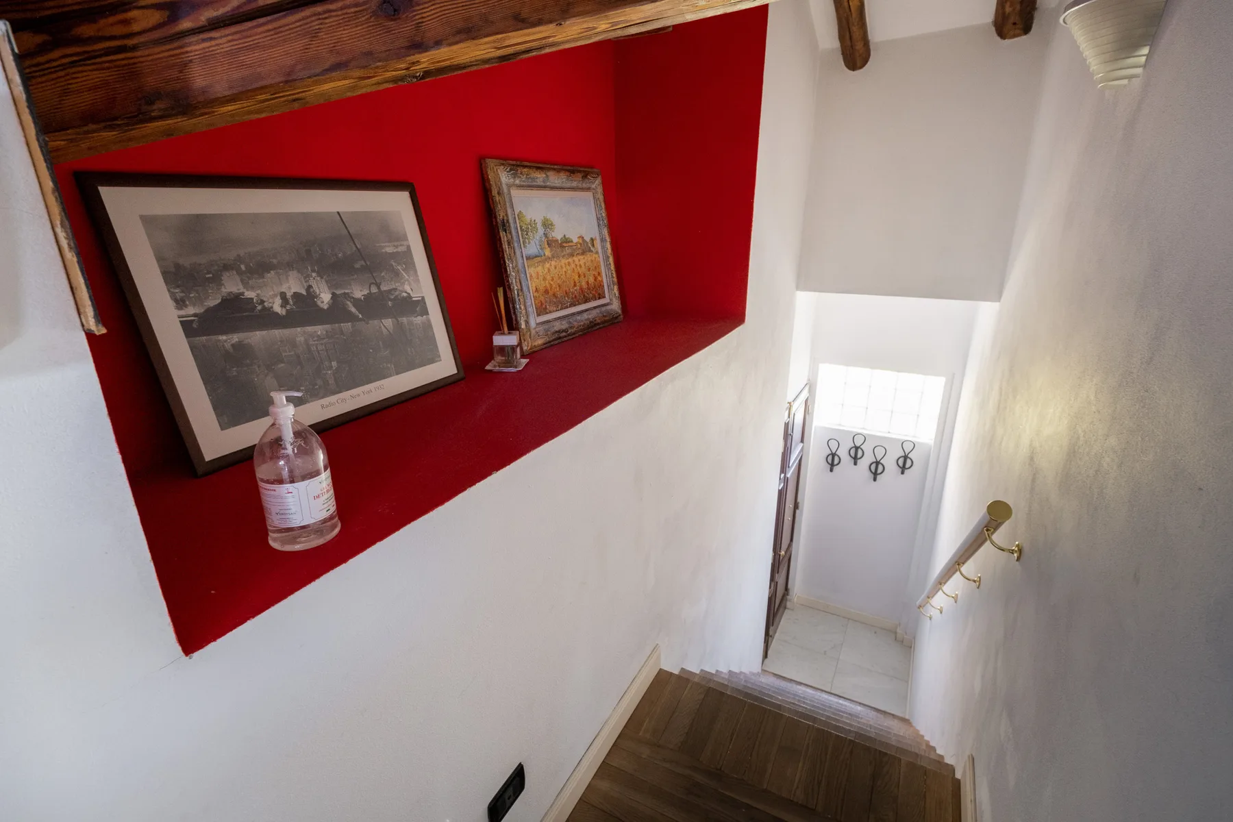 Una escalera estrecha desciende junto a una pared roja con cuadros enmarcados.