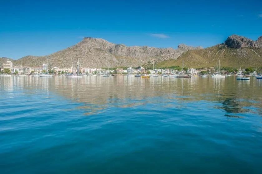 A coastal town and mountain range reflected across calm blue water.