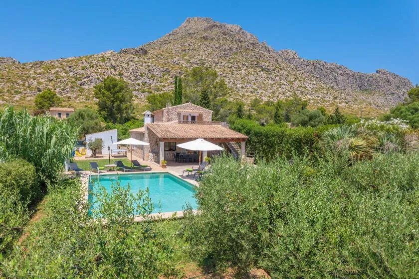 A swimming pool and stone house sit amid greenery below a rocky mountain.