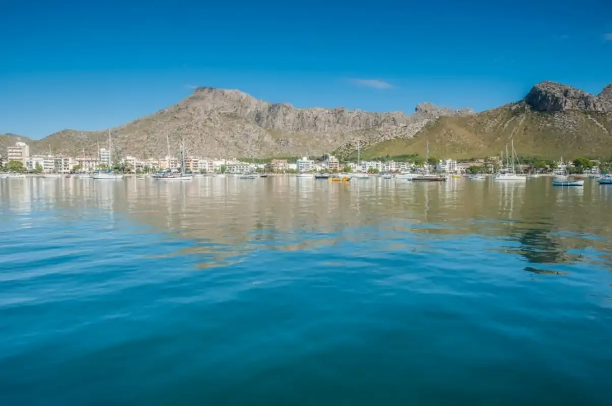 A coastal bay reflects waterfront buildings beneath a rugged mountain range.
