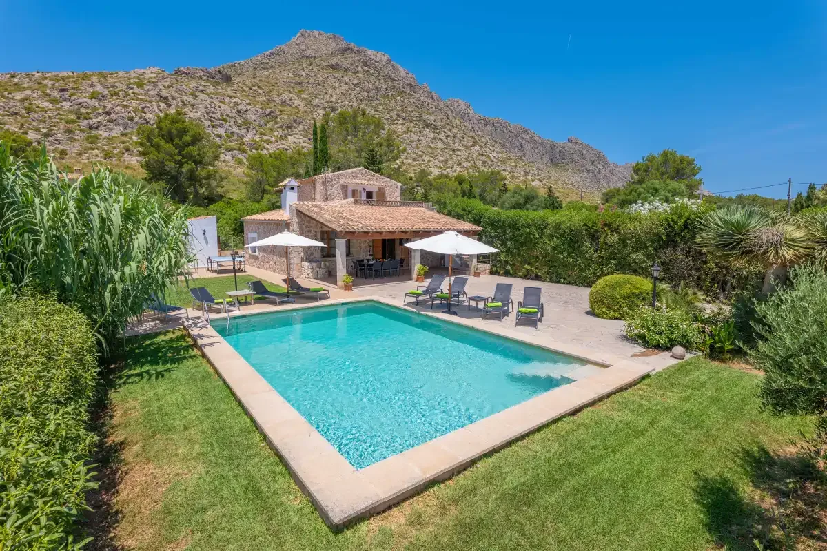 A stone house overlooks a swimming pool and lawn, with a rocky hill behind it.