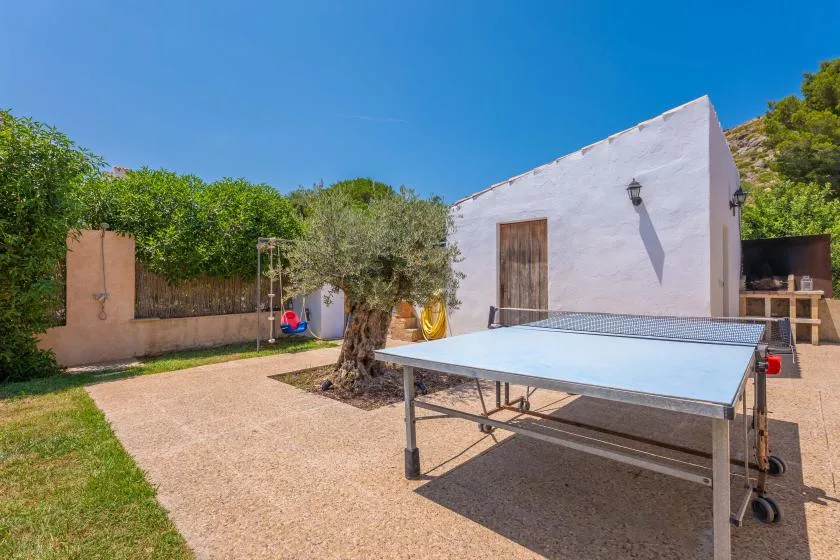 An outdoor table tennis area beside a white building, with trees and greenery.