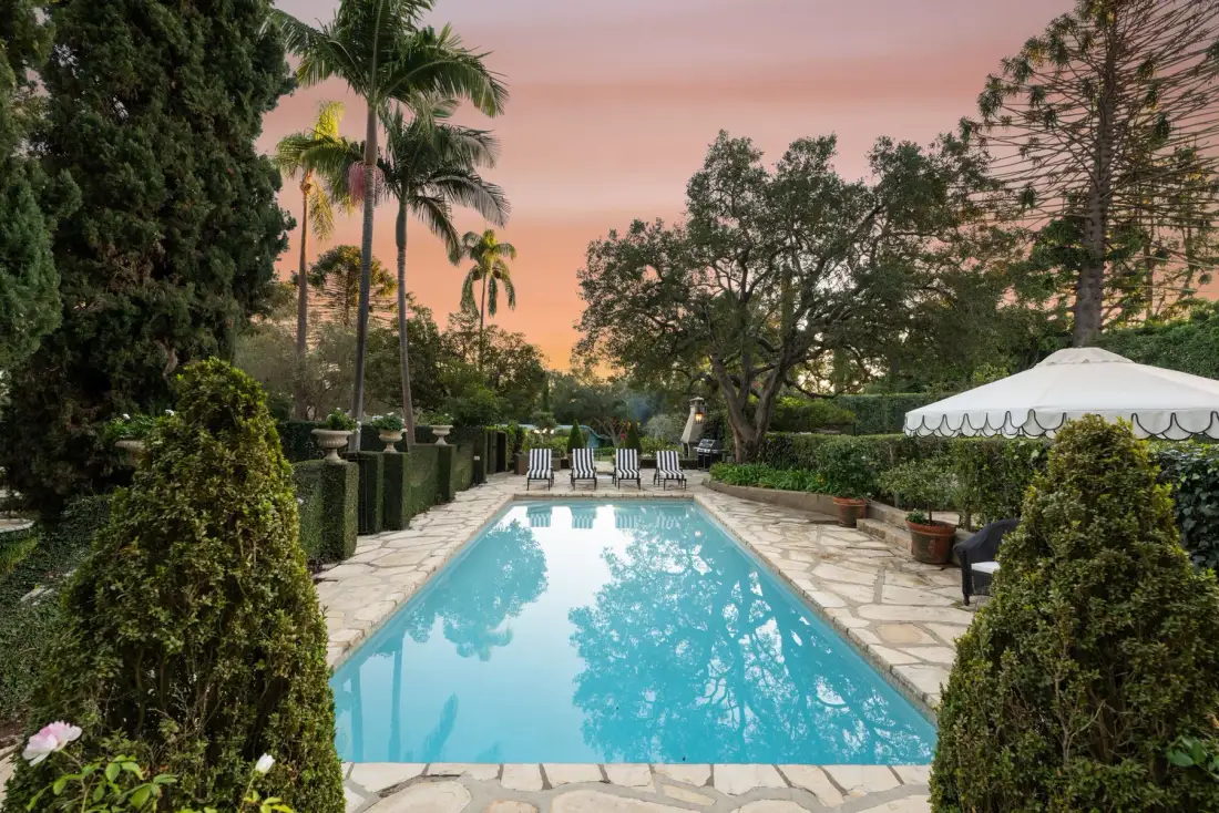 Una piscina de jardín está rodeada de pavimento de piedra y vegetación al atardecer.