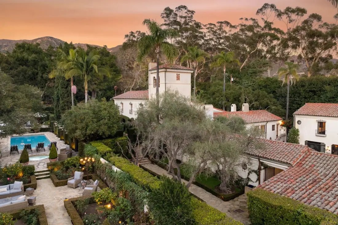 Un patio ajardinado con piscina y edificios blancos bajo un cielo rosado al atardecer.