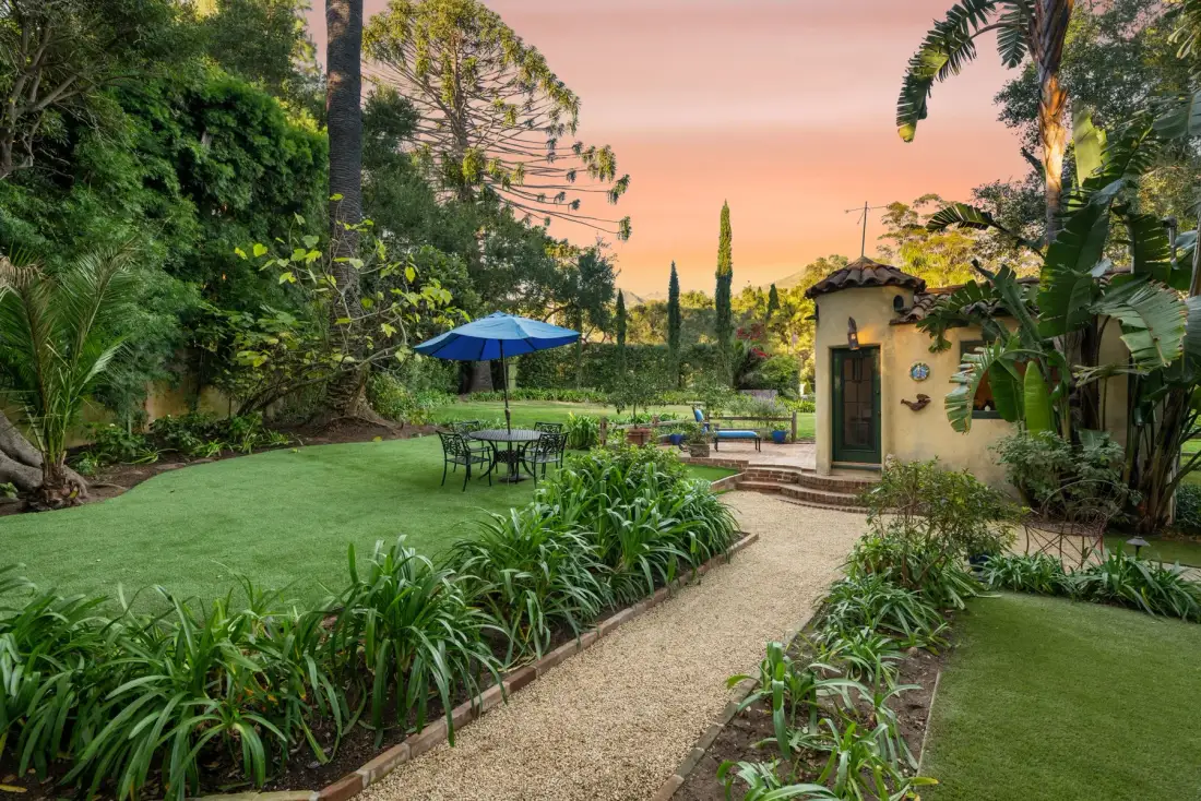 Un jardín ajardinado con un sendero de grava, asientos al aire libre y una pequeña estructura.