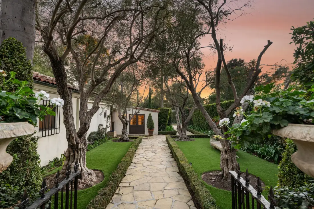 Un sendero de jardín pasa entre céspedes y árboles hacia un edificio blanco al atardecer.