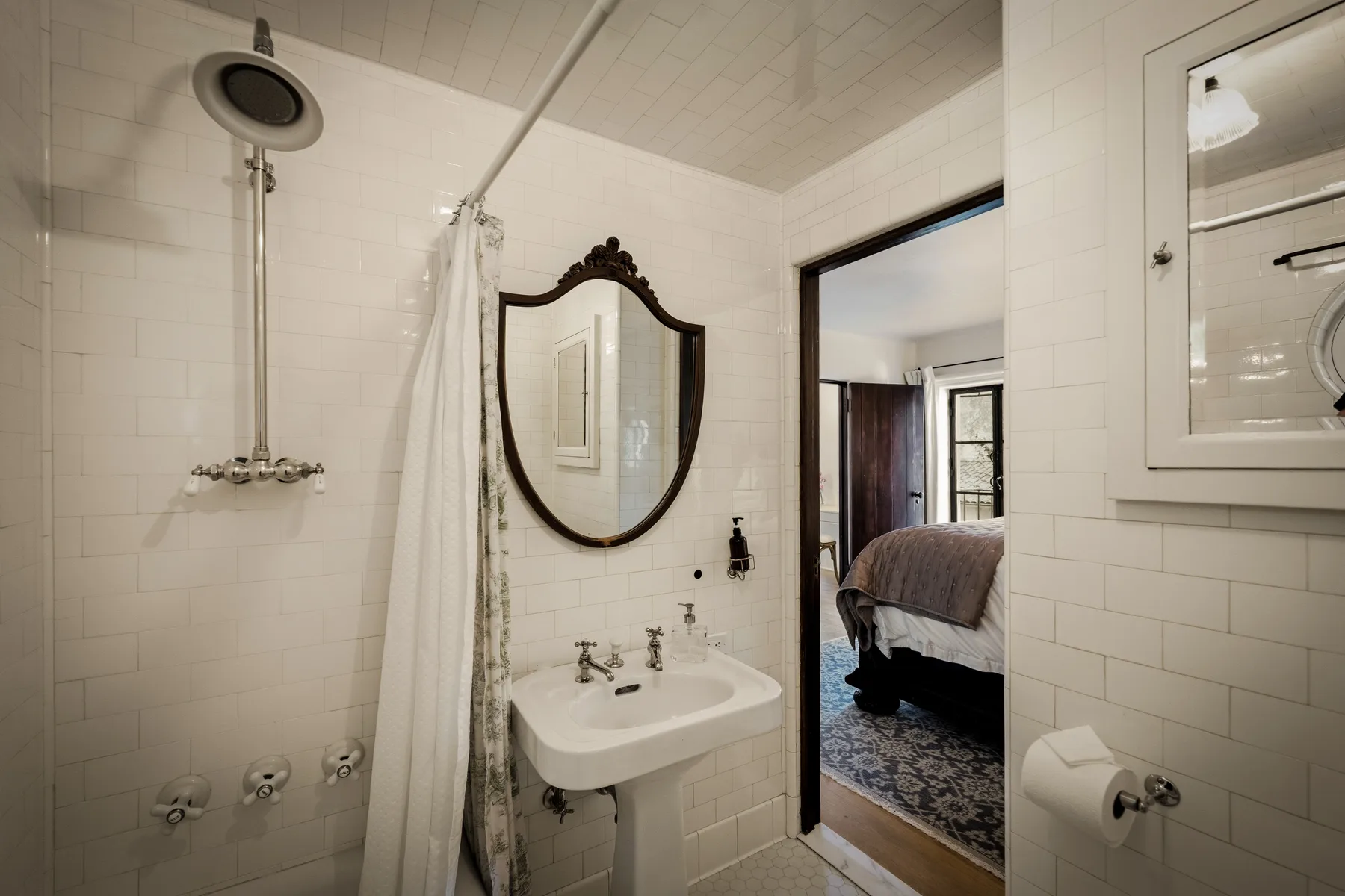 Un baño alicatado con lavabo de pedestal y ducha junto a una puerta abierta.