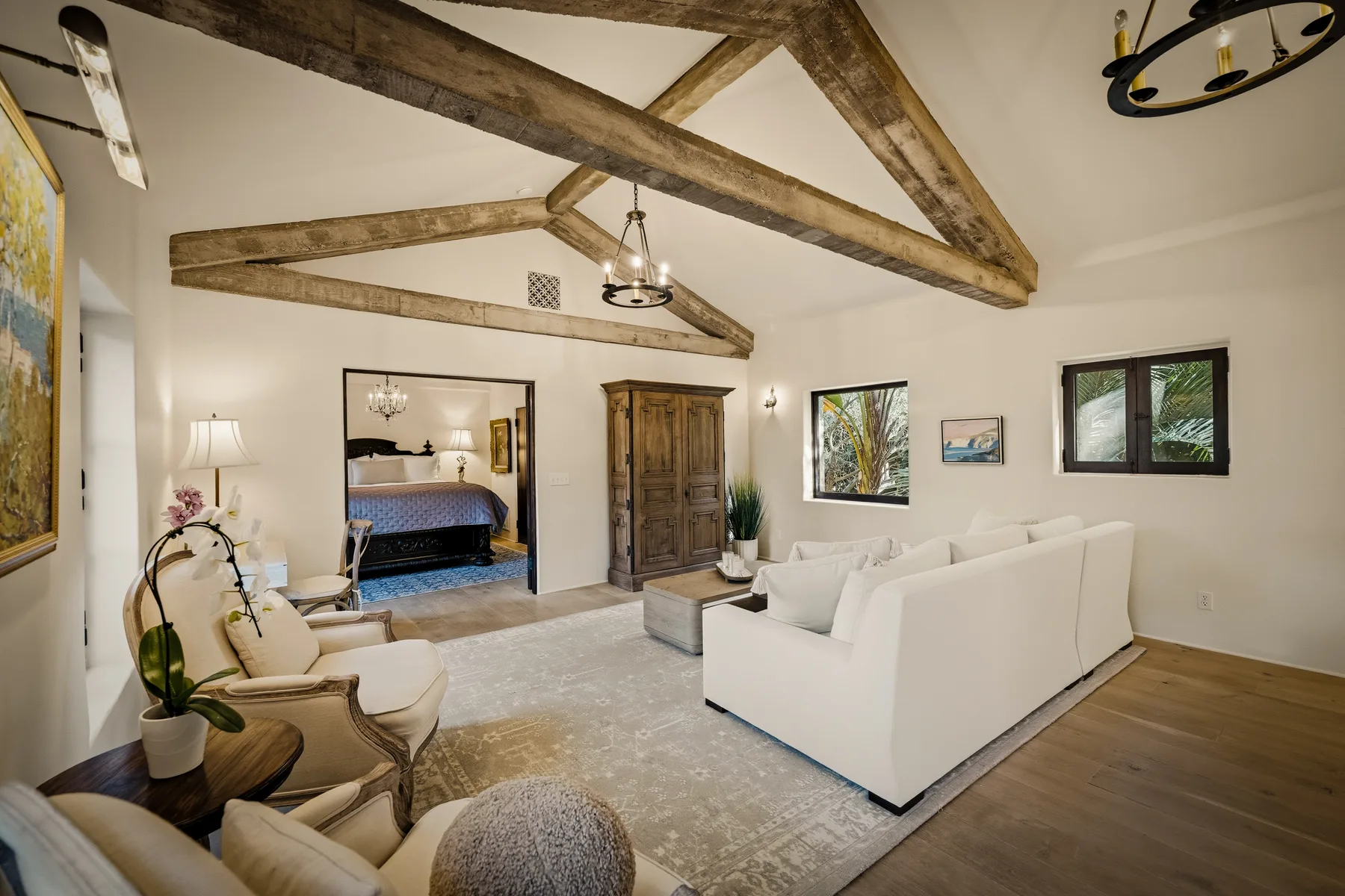 Una sala de estar con asientos blancos, vigas expuestas y vista a un dormitorio.