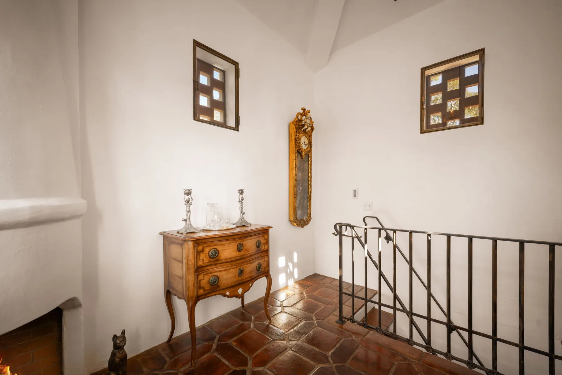 Un rellano interior luminoso con una cómoda de madera, un reloj de pared y una barandilla metálica.