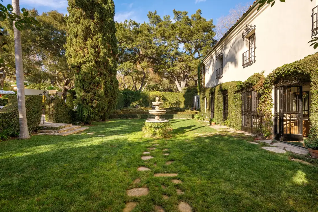 Un jardín ajardinado con una fuente de piedra, árboles y un edificio junto al césped.