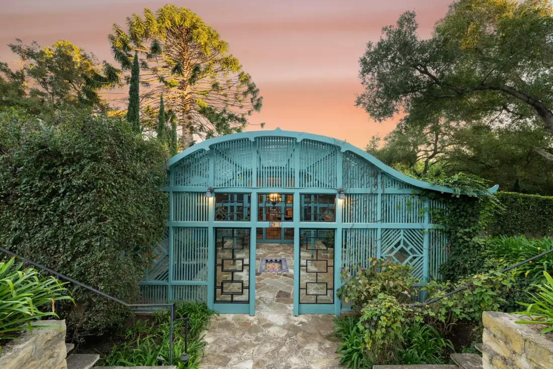 Un pabellón de jardín turquesa con ventanas enrejadas está entre árboles y vegetación.