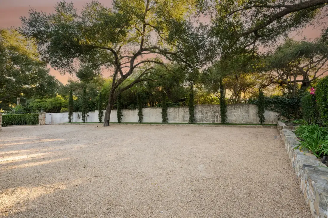 Un patio de grava bajo árboles maduros, bordeado por un muro bajo de piedra.